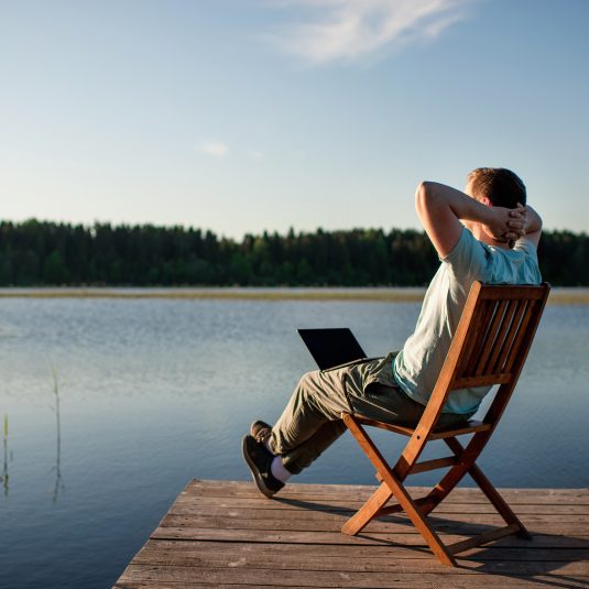 The man is sitting on the chair with the laptop on the lake, working and looking on the setting sun. Concept of remote working.